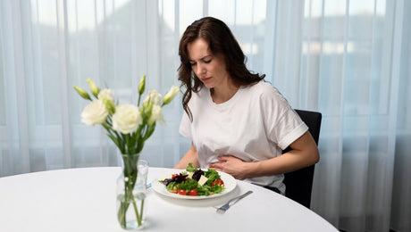 Woman in her 30s sitting at a dining table with a fresh green salad, looking down with discomfort and holding her stomach