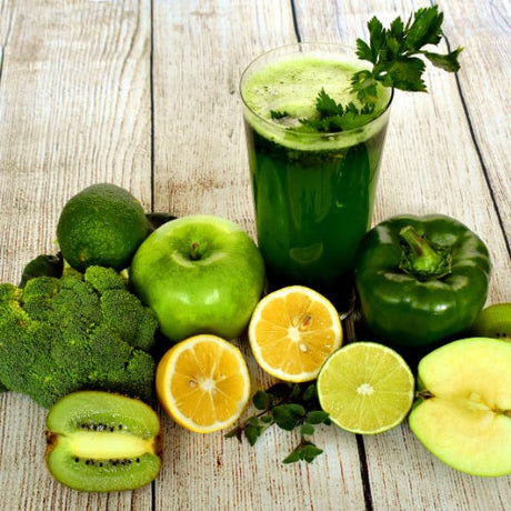 A glass of green juice with apples, lemon, lime, broccoli, bell pepper, and kiwi on a wooden surface.