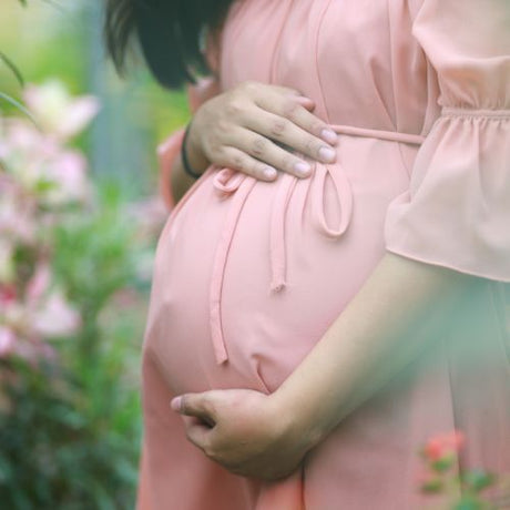 A pregnant woman in a peach dress holds her belly gently while standing among flowers.