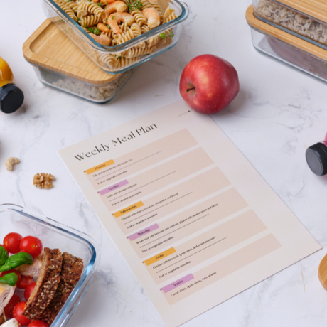 A weekly meal plan sheet on a table with meal prep containers, an apple, and fresh vegetables around it.