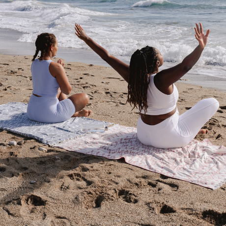 Two women in white activewear practice yoga on beach towels by the ocean, one with arms raised, facing the sea.