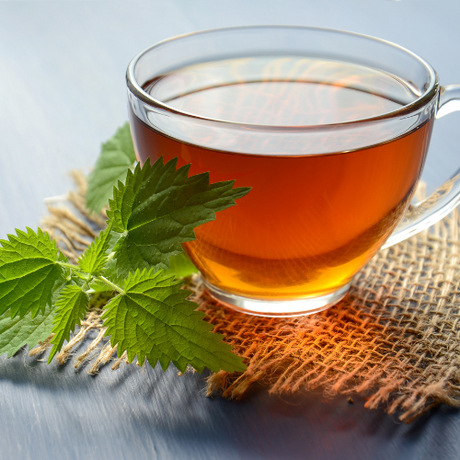 Clear glass cup of herbal tea on a burlap mat, with fresh green nettle leaves beside it.