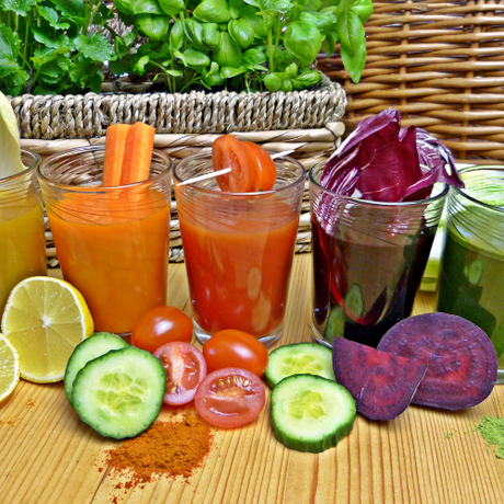Four glasses of colorful vegetable juices with fresh veggies and herbs arranged on a wooden table.