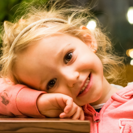 Young child with blonde hair and a big smile, resting her head on her arm while wearing a pink hoodie.