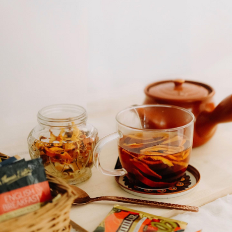 A glass cup of tea with citrus peels, a teapot, and jars of tea on a light surface.