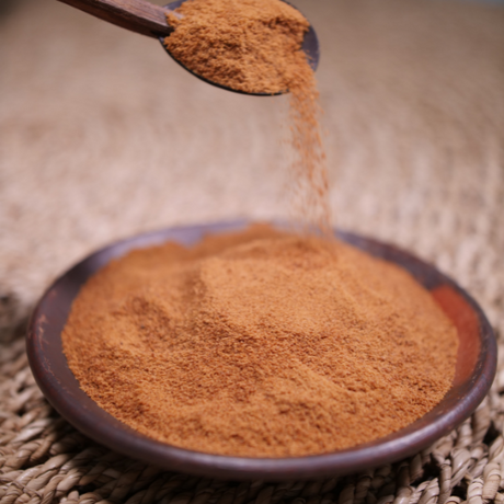 A wooden spoon sprinkles brown powder into a shallow clay bowl filled with the same powder on a woven mat.