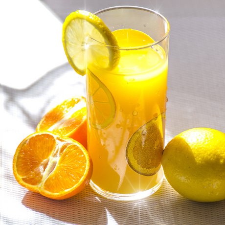 A glass of orange juice with lemon slices, next to fresh orange and lemon halves on a sunlit table.