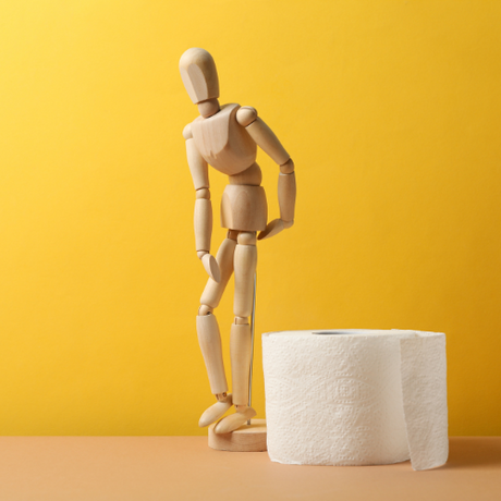 Wooden mannequin leaning forward beside a roll of toilet paper on a yellow background.