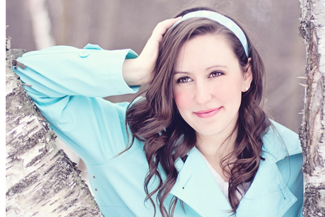 Woman in a light blue coat and headband smiling, standing by birch trees in an outdoor setting.