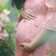 A pregnant woman in a pink dress stands among lush plants, enjoying the calming effects of Pregnancy Tea - 100gms for stress relief.