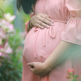 A pregnant woman in a pink dress stands among lush plants, enjoying the calming effects of Pregnancy Tea - 100gms for stress relief.