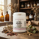 A bottle of Co-Ton Original - 100 Caps rests on a rustic table with dried herbs and glass bottles, emphasizing its herbal formula crafted to help reduce bloating.