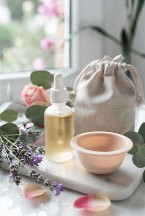 Premium botanical wellness arrangement with a dropper bottle of golden oil, silicone mould, and fabric pouch on marble, accented with lavender, roses, and eucalyptus.